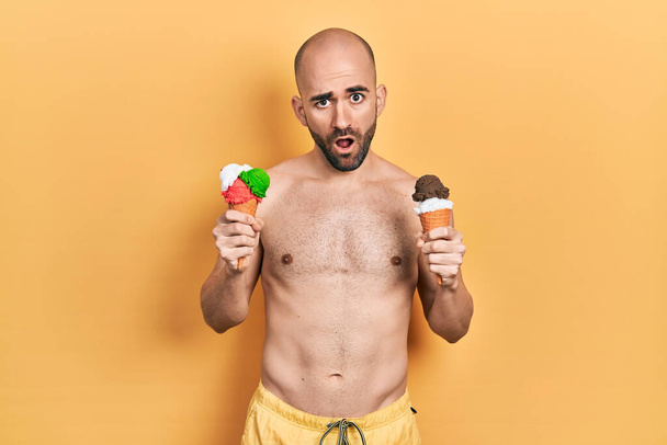 Молодий лисий чоловік у купальнику тримає два конуси морозива в шоковому обличчі, виглядає скептично і саркастично, здивований відкритим ротом
  - Фото, зображення