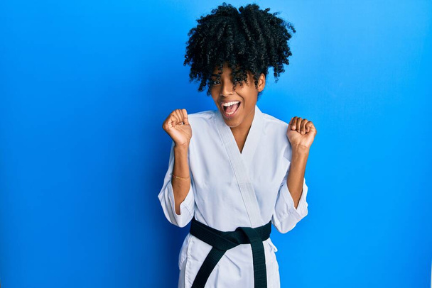 Afro-Amerikaanse vrouw met afrohaar draagt karate kimono en zwarte band vieren verrast en verbaasd voor succes met opgeheven armen en open ogen. winnaarsconcept.  - Foto, afbeelding