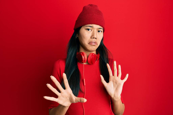 Młoda Chinka słuchająca muzyki za pomocą słuchawek brzydziła się ekspresją, niezadowolona i przerażona robiąc obrzydliwą minę, bo reakcja awersji. z podniesionymi rękami  - Zdjęcie, obraz