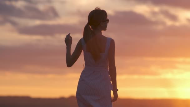Темный силуэт молодой женщины в летнем платье, стоящей на открытом воздухе и наслаждающейся видом на природу на закате. - Кадры, видео