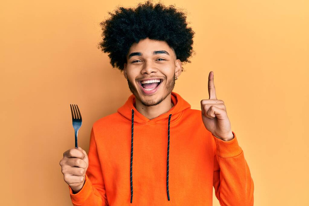 Νεαρός Αφροαμερικάνος με αφρο μαλλιά κρατώντας ένα ασημένιο πιρούνι χαμογελώντας με μια ιδέα ή ερώτηση δείχνοντας το δάχτυλο με χαρούμενο πρόσωπο, νούμερο ένα  - Φωτογραφία, εικόνα