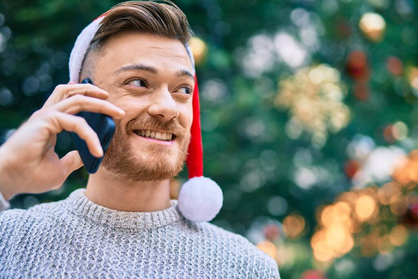 Νεαρός καυκάσιος που φοράει χριστουγεννιάτικο καπέλο και μιλάει στο κινητό του στο πάρκο - Φωτογραφία, εικόνα