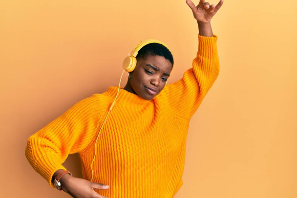 Krásná africká americká mladá žena s krátkými vlasy nosí sluchátka poslech hudby a tanec přes izolované žluté pozadí - Fotografie, Obrázek