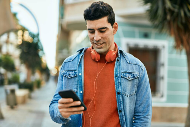 Mladý hispánec se šťastně usmívá pomocí smartphonu a sluchátek ve městě. - Fotografie, Obrázek
