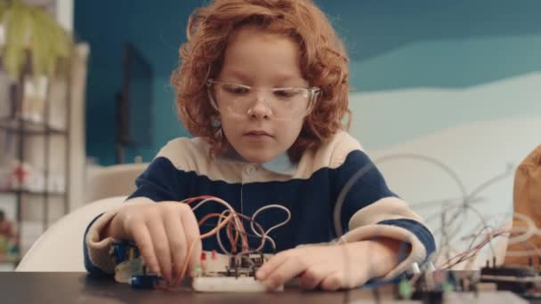 Közepes felvétel kaukázusi vörös hajú iskolás fiú visel alkalmi ruhák ül asztali az osztályteremben, és építő robot segítségével műanyag részletek és vezetékek - Felvétel, videó