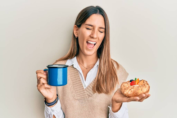 Brunette young woman drinking coffee and eating pastry smiling and laughing hard out loud because funny crazy joke.  - Photo, Image