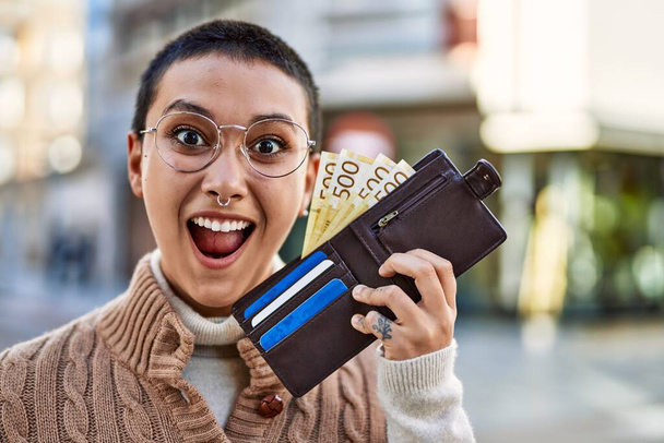Schöne hispanische Frau mit kurzen Haaren lächelt glücklich im Freien und hält Brieftasche mit norwegischen Banknoten - Foto, Bild