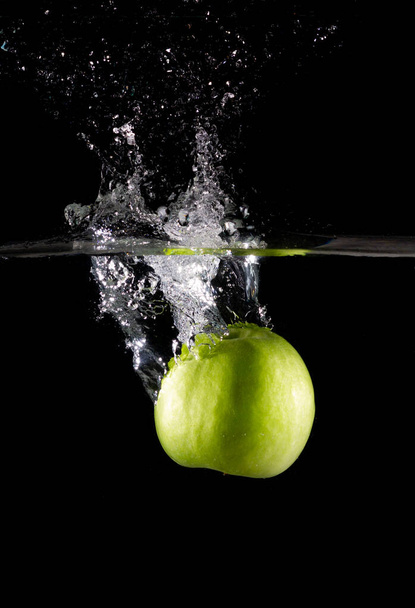 A vertical shot of a green apple in water with splashes on a dark isolated background - Zdjęcie, obraz