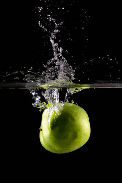 A vertical shot of a green apple in water with splashes on a dark isolated background - 写真・画像