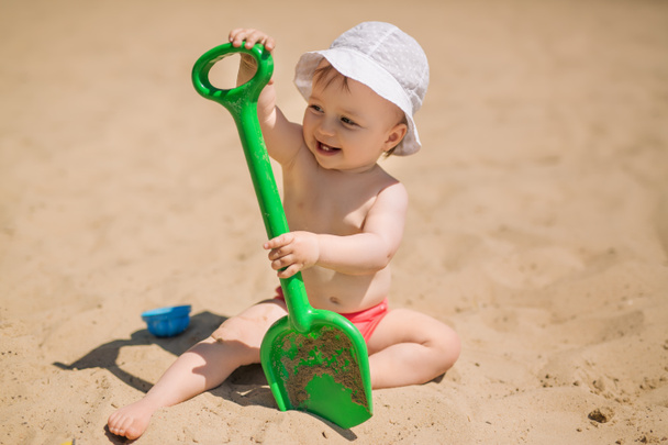 Sıcak bir günde kumsalda büyük yeşil oyuncak küreği olan bir çocuk. Çocuklar için güneş kremi. Panama şapkalı ve külotlu bir çocuk kumlarda oynuyor. - Fotoğraf, Görsel