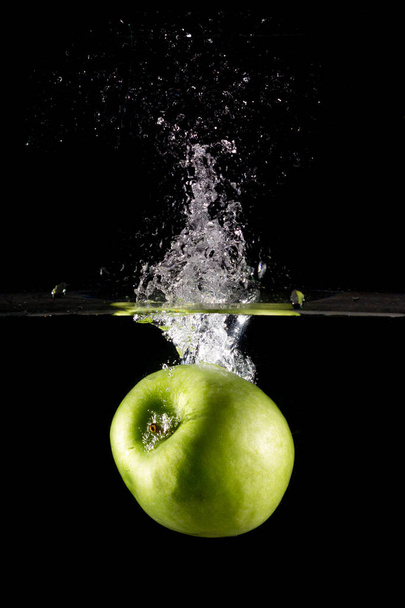 A vertical shot of a green apple in water with splashes on a dark isolated background - Фото, зображення