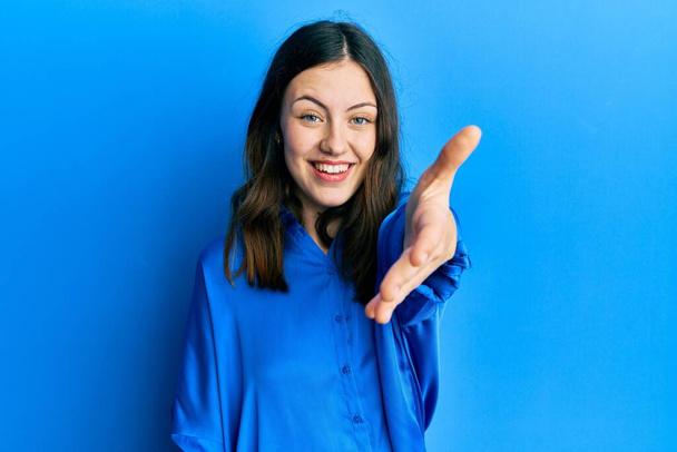 Mujer morena joven con camisa azul casual sonriente amistoso ofreciendo apretón de manos como saludo y bienvenida. negocios exitosos.  - Foto, Imagen