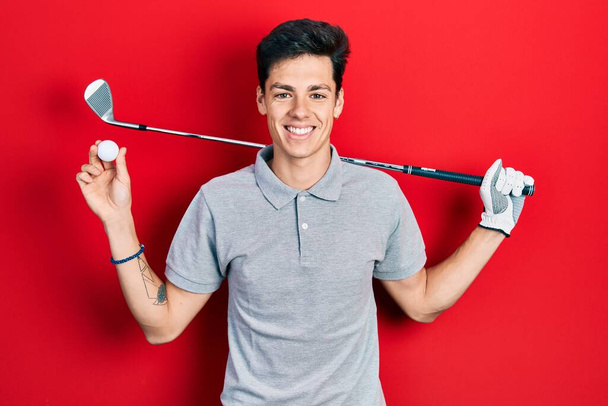 Junger hispanischer Mann mit Golfball, der mit einem fröhlichen und kühlen Lächeln im Gesicht lächelt. Zähne zeigen.  - Foto, Bild