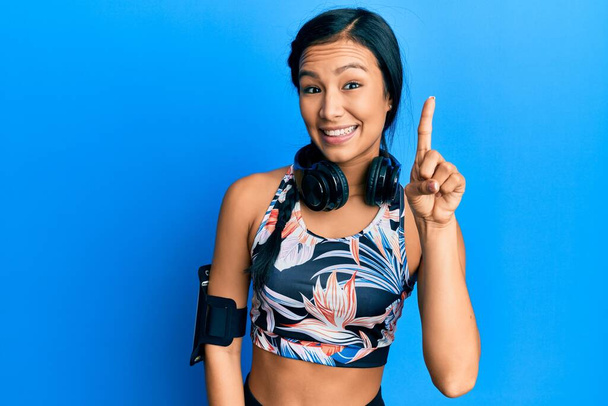Beautiful hispanic woman wearing gym clothes and using headphones smiling with an idea or question pointing finger up with happy face, number one  - Foto, imagen