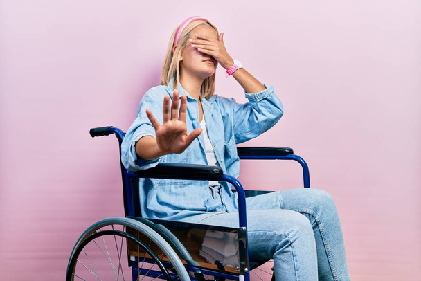 Красивая блондинка, сидящая на инвалидной коляске, закрывая глаза руками и делая стоп-жест с грустным и испуганным выражением. смущенная и негативная концепция.  - Фото, изображение