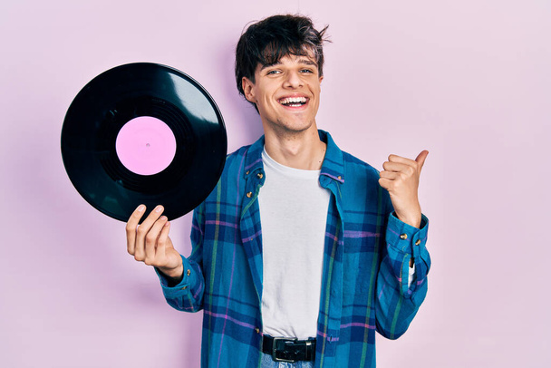 Fešák hipster mladý muž drží vinyl disk ukazující palec nahoru na stranu usmívá šťastný s otevřenými ústy  - Fotografie, Obrázek