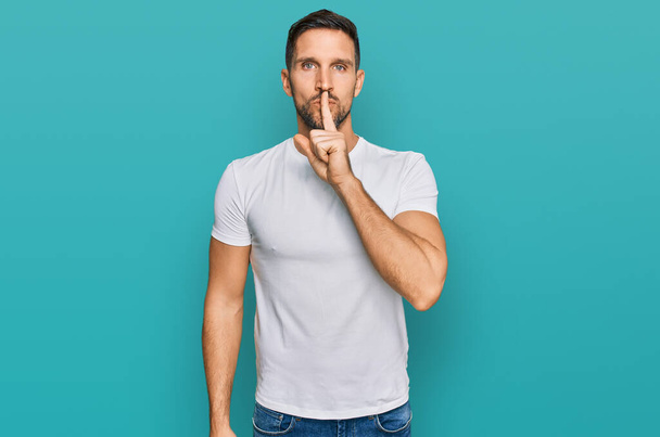Un hombre guapo con barba que usa una camiseta blanca y casual pidiendo silencio con el dedo en los labios. silencio y concepto secreto.  - Foto, Imagen