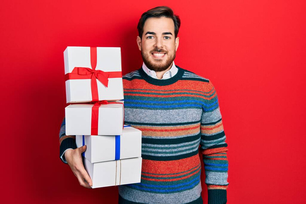 Hombre caucásico guapo con barba sosteniendo regalos que parecen positivos y felices de pie y sonriendo con una sonrisa confiada mostrando los dientes  - Foto, imagen