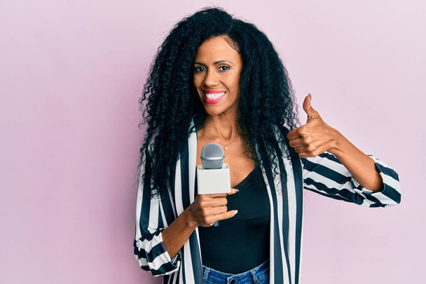 Moyen âge femme afro-américaine tenant microphone journaliste souriant heureux et positif, pouce levé faire excellent et signe d'approbation  - Photo, image