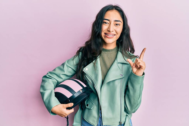latino tiener meisje met gebit beugels houden motorfiets helm glimlachen gelukkig wijzend met hand en vinger naar de zijkant  - Foto, afbeelding