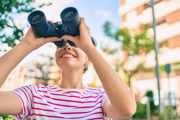 Joven chica hispana sonriendo feliz buscando una nueva oportunidad usando prismáticos en la ciudad. - Foto, Imagen