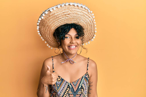 Mujer hispana joven sosteniendo sombrero mexicano sonriendo feliz y positivo, pulgar hacia arriba haciendo excelente y signo de aprobación  - Foto, imagen