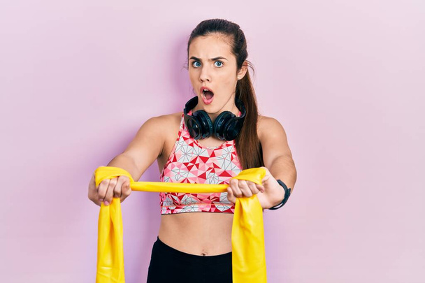 Young brunette teenager training arm resistance with elastic arm bands afraid and shocked with surprise and amazed expression, fear and excited face.  - Photo, Image