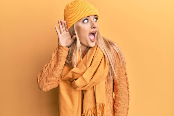 Mulher caucasiana jovem vestindo camisola de inverno de lã e boné sorrindo com a mão sobre o ouvido ouvindo um rumor ou fofoca. conceito de surdez.  - Foto, Imagem