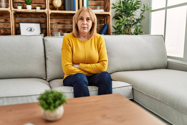Keski-ikäinen valkoihoinen nainen päällään rento vaatteita istuu sohvalla kotona skeptinen ja hermostunut, paheksuva ilme kasvot ristissä kädet. negatiivinen henkilö.  - Valokuva, kuva
