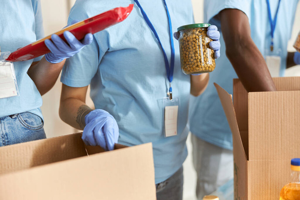 Close up of volunteer in protective gloves holding jar of peas while sorting and packing foodstuff in cardboard box, working on donation project together with team - Photo, Image