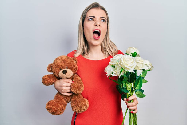 Belle femme caucasienne tenant ours et bouquet de fleurs pour anniversaire en colère et fou hurlant frustré et furieux, criant de colère. rage et concept agressif.  - Photo, image