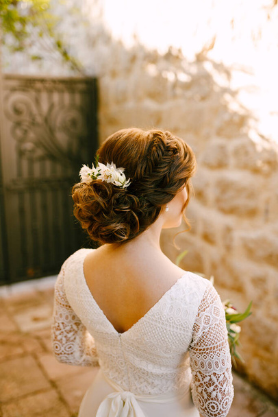 Κομψή νύφη στέκεται πίσω σε ένα λευκό φόρεμα δαντέλα με ένα πολυτελές χτένισμα και λουλούδια στα μαλλιά της κοιτάζει στον τοίχο απέναντι - Φωτογραφία, εικόνα