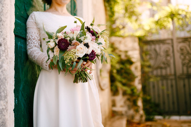 Panna młoda trzyma w rękach piękny bukiet kwiatów opierając się o zielone drzwi budynku - Zdjęcie, obraz