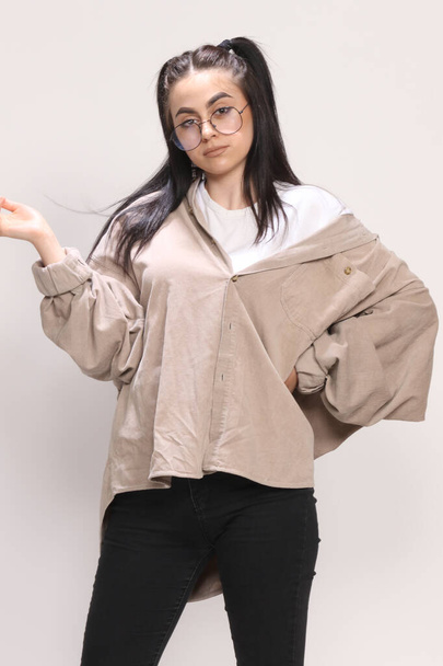 Beautiful caucasian hipster girl with black hair and glasses is posing in studio on isolated background. Style, trends, fashion concept. - Photo, Image