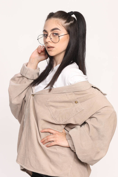 Beautiful caucasian hipster girl with black hair and glasses is posing in studio on isolated background. Style, trends, fashion concept. - Valokuva, kuva