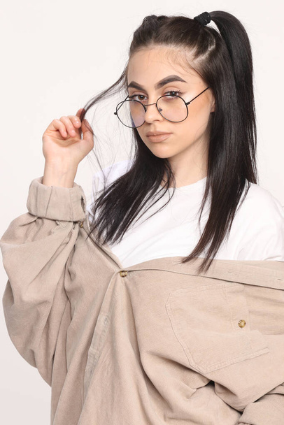 Hermosa chica hipster caucásica con el pelo negro y gafas está posando en el estudio sobre fondo aislado. Estilo, tendencias, concepto de moda. - Foto, Imagen