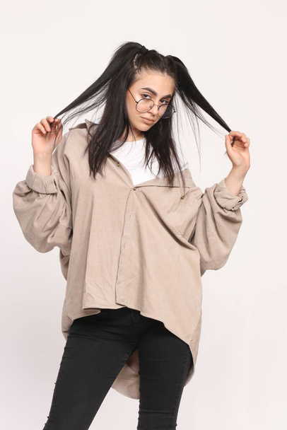 Beautiful caucasian hipster girl with black hair and glasses is posing in studio on isolated background. Style, trends, fashion concept. - 写真・画像