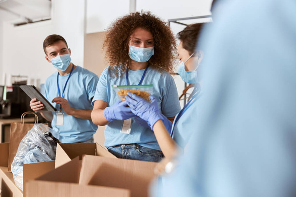 Drukke vrijwilligers in blauw uniform, beschermende maskers en handschoenen die een pakket pasta 's en andere voedselwaren sorteren tijdens hun werk in liefdadigheidsinstellingen - Foto, afbeelding