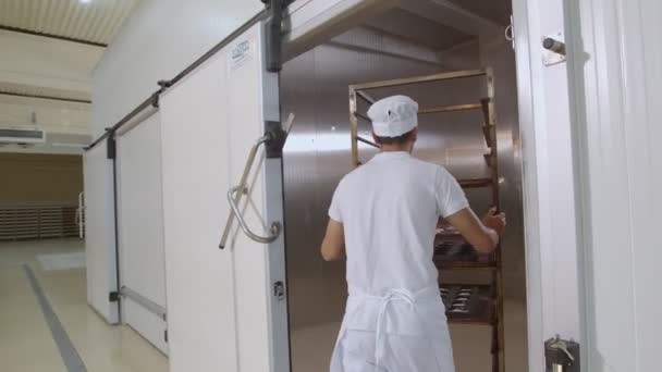 La macchina fotografica segue il panettiere vestito di bianco e toque dal retro mentre mette un carrello con pane pronto da cuocere nel grande forno - Filmati, video