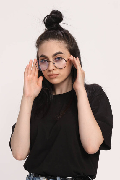 Beautiful caucasian hipster girl with black hair and glasses is posing in studio on isolated background. Style, trends, fashion concept. - 写真・画像