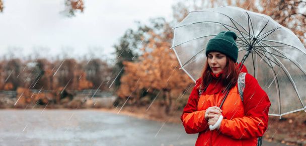 Krásná dívka drží transparentní deštník během deště v podzimním oblačném počasí na podzim. Žena v červené nepromokavé podzimní bundě ve městě - Fotografie, Obrázek