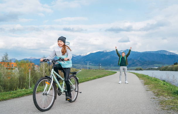 Smiling kids on a bicycle asphalt path with snowy mountains background. Brother helping to sister. He rose arms up after a successful riding start. Happy childhood concept image. - Zdjęcie, obraz