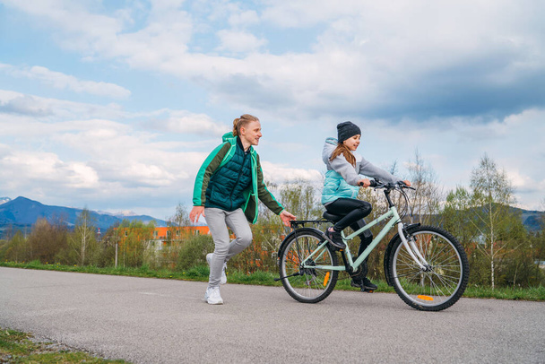 Hymyilevät lapset polkupyörällä asfalttitiellä. Veli auttaa siskoa ja opettaa tekemään ensimmäiset askeleet ratsastus. Happy lapsuuden käsite kuva. - Valokuva, kuva