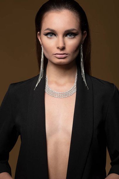beautiful young woman with diamond jewellery , on dark beige background , she is wearing a black jacket , shining necklace and earrings,  - Foto, Imagem