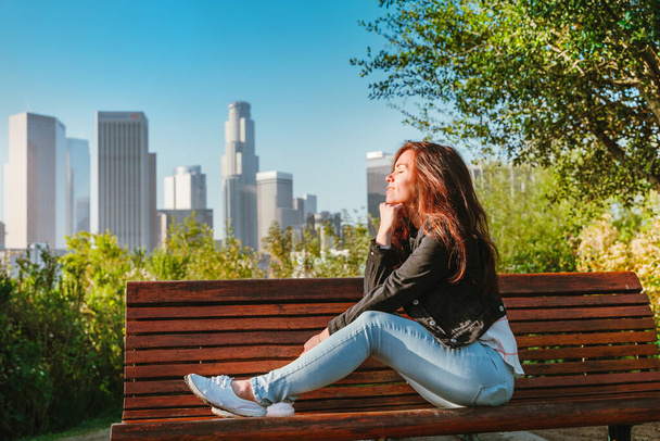 Impresionante mujer morena en una chaqueta de mezclilla se sienta en un banco del parque con vistas a los rascacielos del centro de Los Ángeles - Foto, Imagen