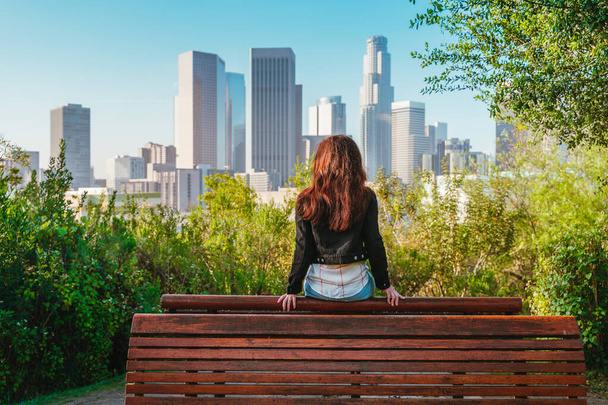 Stunning brunette woman in a denim jacket sits on a park bench with a view of Downtown skyscrapers in Los Angeles - Photo, Image