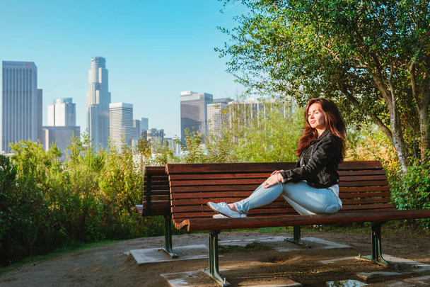 Потрясающая брюнетка в джинсовой куртке сидит на скамейке в парке с видом на небоскребы в центре Лос-Анджелеса - Фото, изображение