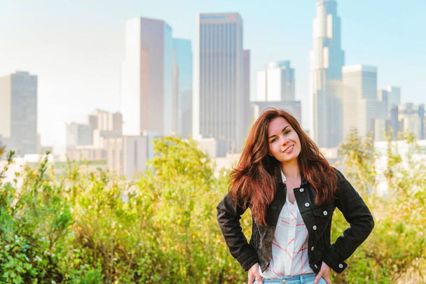 Stunning brunette woman in a denim jacket sits on a park bench with a view of Downtown skyscrapers in Los Angeles - Photo, Image