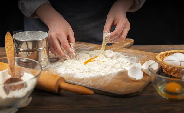 Women's hands, flour and dough. A woman, in an apron prepares dough for homemade baking, a rustic home cozy atmosphere, a dark background with unusual lighting. - 写真・画像
