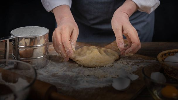 Women's hands, flour and dough. A woman, in an apron cooking dough for homemade baking, a rustic home cozy atmosphere, a dark background with unusual lighting. - Fotoğraf, Görsel
